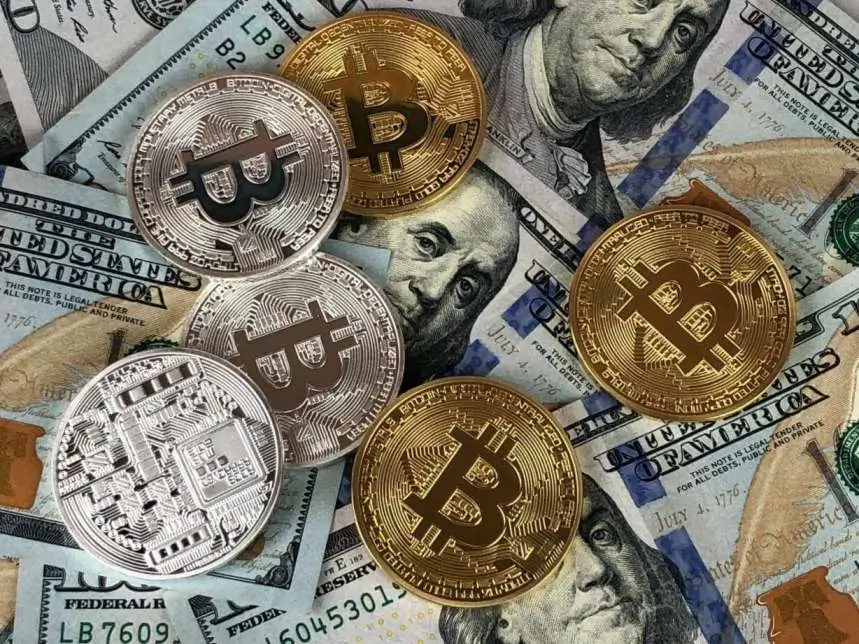 A picture of paper currency lying behind a handful of gold and silver coins with cryptocurrency symbols like Bitcoin. Cryptocurrency is a growing method for storing digital assets, consumer purchases, and long-term investment.